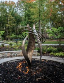 A modern sculpture with intertwined metal shapes stands surrounded by a fire pit, set in a landscaped garden with a backdrop of trees and foliage.