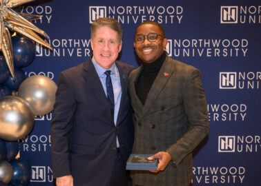 Two men in suits stand together smiling in front of a Northwood University backdrop, with one holding a plaque.