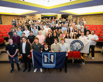 A large group of students gathers in a lecture hall, proudly holding a Northwood University flag and a university seal flag, united by their mission to stay connected.
