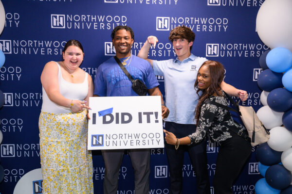 Four individuals pose in front of a Northwood University backdrop. One person holds a sign that reads "I DID IT!" Blue and white balloons are on the right side, celebrating their achievements and encouraging everyone to stay connected.