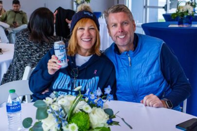Two people sitting at a table with drinks, smiling at the camera. One holds a can and wears a "Northwood" sweatshirt, while the other wears a blue vest. In the foreground is a flower arrangement. It’s clear they cherish these moments to stay connected.