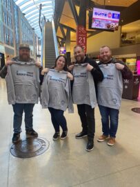 Four people holding Detroit Basketball sweatshirts pose inside a modern building with glass walls and stairs in the background, showcasing how fans can stay connected to their team.