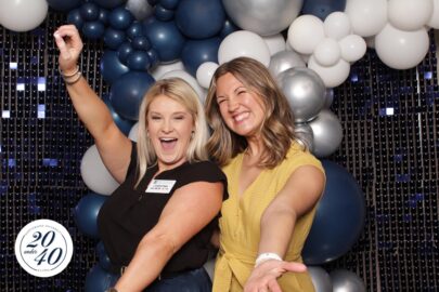 Two women pose enthusiastically in front of a backdrop with blue and white balloons. One is wearing a black sleeveless top with a name tag, and the other is in a yellow top. A "20 under 40" logo appears, encouraging young professionals to stay connected.
